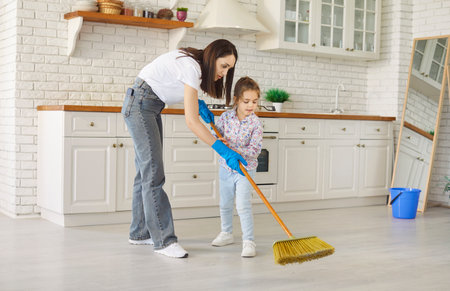 Loving mother and little helper daughter learning sweeping the floor together with broom wearing blue cleaning gloves in bright, spacious modern kitchen area room, doing daily household choresの写真素材