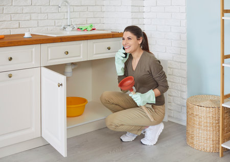 Young smiling girl talking by phone advising with plumber how to unclog a sink drain in kitchen. Woman trying to clean sink using plunger at home. Housekeeping and household work conceptの写真素材