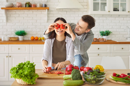 Happy funny young family couple, woman, man healthy vegans at home kitchen, vegetarian diet, vegetables, plate of fresh eco food at table, health benefits of well-balanced food, tomato eyes showingの写真素材