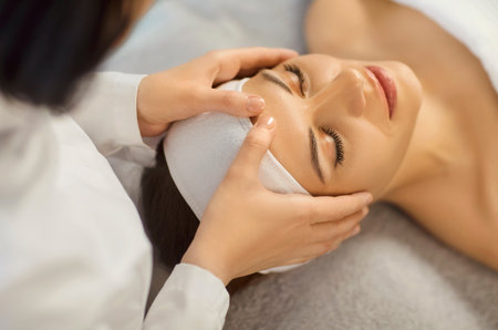 Close up of young woman enjoying relaxing face massage at beauty spa salon. Female client lying with eyes closed while esthetician gently massaging skin during professional facial skincare treatment.の写真素材