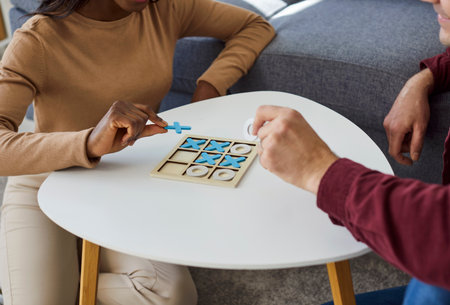 Cropped closeup portrait of young multinational couple having fun playing tic tac toe game together at home on weekend. People friends playing board game in living room. Hobbies and leisure concept.の写真素材