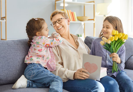 Happy smiling children congratulating grandmother with Women Day or Mothers day, greeting with card and bouquet of yellow tulips, posing on sofa in living room. Family and celebration conceptの写真素材
