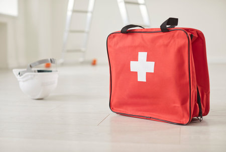 Closeup of red bag with first aid kit standing in empty building site with safety helmet and construction ladder on floor. Emergency, health insurance and job accident at work concept.の写真素材