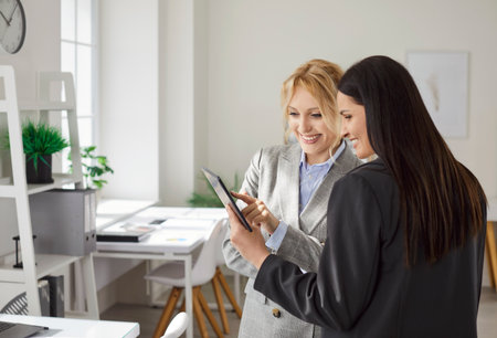 Portrait of young cheerful female business people employees standing on workplace, looking at tablet monitor screen and discussing job project or company finance. Two women working in office.の写真素材