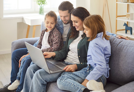 Happy young family sitting together on sofa and looking at laptop screen and watching something fun. Smiling parents and two children hugging, using laptop and spending quality time together at home.の写真素材