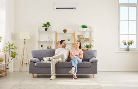 Happy couple sitting on comfortable gray sofa in living room, relaxing together under air conditioner. Smiling young man and woman resting on couch, talking and enjoying fresh cool air at home.の写真素材