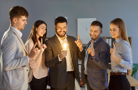 Smiling group of business people standing in dark office with index fingers up and looking at glowing light bulb. Happy colleagues generating new creative ideas and innovations and sharing inspirationの写真素材