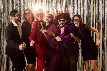 Portrait of happy excited young people group in bright suits enjoying disco corporate party together and having fun on silver sparkle foil background. Dancers in night club. Celebration concept.の写真素材