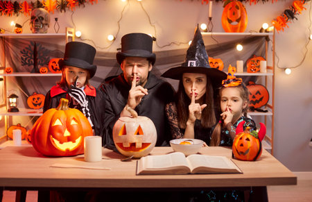 Portrait of family of mother, father and two children in masquerade costumes preparing for Halloween party at home saying hush be quiet with fingers on lips with shhh gesture. Celebration concept.の写真素材