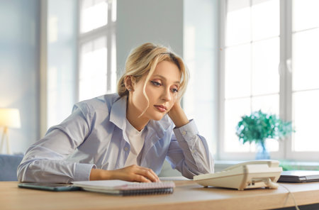 Tired young woman sitting at desk, leaning on hand and staring at landline phone. Exhausted anxious company employee waiting for important phone call, feeling stressed and tense while expecting news.の写真素材