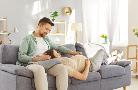 Young man sitting on sofa in living room at home talking with pregnant wife touching her big belly. Happy couple expecting a baby preparing to become parents. Pregnancy and parenthood concept.の写真素材