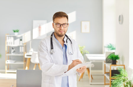Portrait of focused doctor in white coat standing in medical office and writing notes on clipboard. Serious healthcare professional with glasses and stethoscope looking at camera. Health care concept.の写真素材