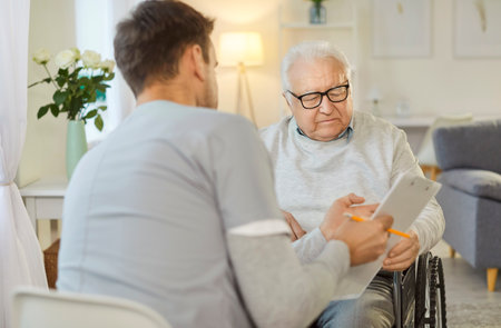 Male nurse sitting back to camera, holding tablet, showing results of analysis to handicapped senior man in wheelchair, explains prescription, takes care of him while being on rehabilitationの写真素材