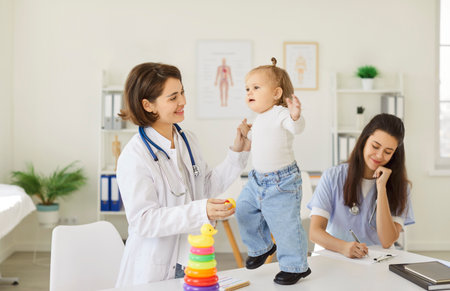 Smiling female pediatrician playing with toddler standing on desk during medical checkup. Friendly caring doctor holding toy to engage little child while nurse taking notes after examination in clinicの写真素材