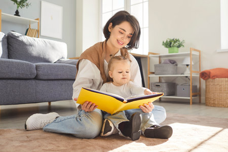 Smiling young mother sitting on floor with her little child in bright living room, reading fairytale book together. Caring mom and toddler baby learning, enjoying quality time. Family leisure concept.の写真素材
