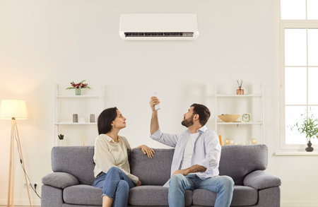Young family couple sitting together on sofa in living room and turning on air conditioner. Focused relaxed man and woman adjusting settings with remote controller for comfortable temperature indoor.の写真素材