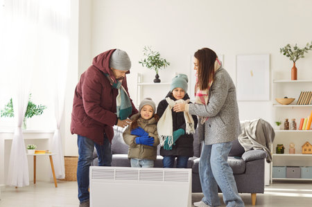 Young freezing family with two children in chilly living room wearing winter outwear trying to warm up standing near electric heater getting cold at home. Heating problems concept.の写真素材