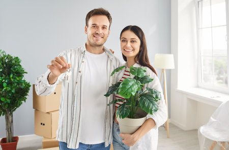 Portrait of young cheerful couple standing in bright room of new home surrounded by cardboard boxes and showing keys to camera. Happy smiling family holding potted flower and hugging on moving day.の写真素材