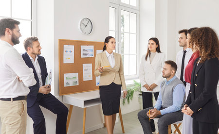 Group of young business people on meeting discussing finance and work project standing in office. Company employees listening confident woman colleague with a report in conference room.の写真素材