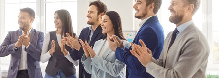 A group of six smiling professionals is clapping in a modern office setting. The diverse team showcases a sense of achievement during a corporate event.の写真素材