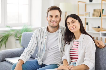 Portrait of young happy married couple sitting on sofa in living room at home. Smiling family man and woman on couch looking cheerful at camera. Harmonic relationships and Valentines day concept.の写真素材