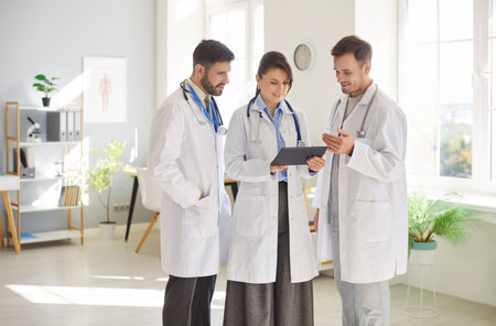 Portrait of a team of three young cheerful doctors in white uniform looking at tablet screen discussing treatment standing in hospital or clinic. Medical staff and health care concept.の写真素材