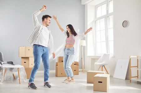 Young couple celebrating moving day smiling and dancing in new apartment full of cardboard boxes. Family having fun happy to buy a property. Move, relocating, real estate, mortgage concept.の写真素材