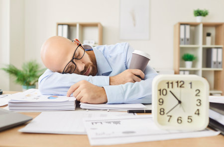 Tired bald man sleeping on a table surrounded by office documents while holding a coffee cup and...