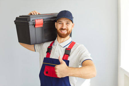 Portrait of a happy smiling repairman holding tool box in hands in empty room looking cheerful at camera and showing thumb up sign. Professional young handyman worker standing indoors in blue overallの写真素材