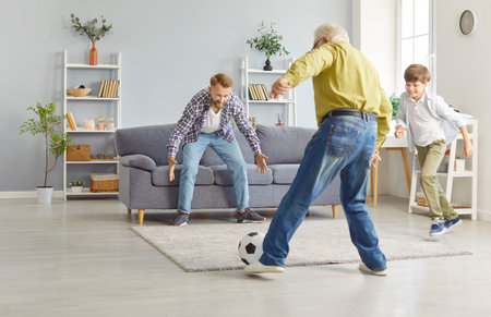 Happy family playing football. In bright living room, three generations of men are having fun playing and passing ball. Lively and nimble grandfather kicks ball towards his son and little grandson.の写真素材