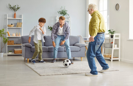Happy little child boy playing football game with elderly senior grandfather and young smiling father in the living room at home enjoying spending time together. Family leisure concept.の写真素材