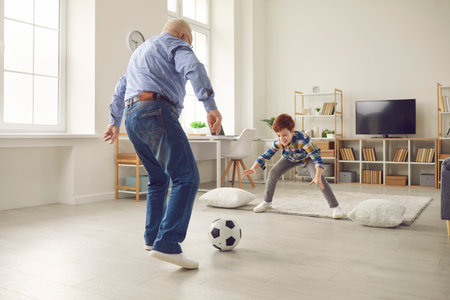 Grandfather and a boy are playing football together at home, enjoying active leisure time. Family bonding moment involves kicking a ball, highlighting shared activity and connection through the game.の写真素材