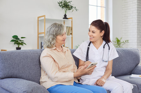 Young cheerful female caregiver teaching senior person to handle with mobile phone at home. Friendly nurse showing elderly woman how to use smartphone. Nursing home service and patient support.の写真素材