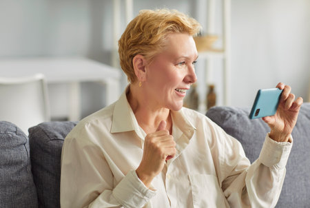 Senior mature woman looking at mobile phone having video call sitting on sofa in living room at home. Female person talking online with smartphone. Retirement and communication technology.の写真素材