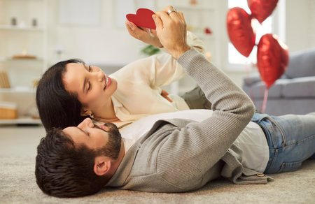 Happy family couple celebrating Valentines Day at home. Smiling young man and woman lying on floor in living room looking at red heart shaped card in hands. Love, romance, holiday conceptの写真素材