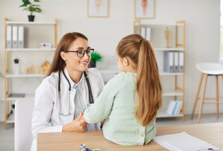 Back view of little preschool girl sitting on desk and holding hands with friendly family doctor during examination in hospital. Smiling female pediatrician cheering up child patient during checkup.の写真素材
