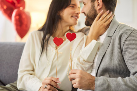 Smiley loving couple celebrates Valentines Day with a kiss and warm embrace, holding red heart shapes. Affection, love, and the joyful essence of the holiday celebration and romance.の写真素材