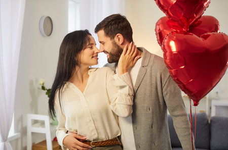Young happy man holding red foil balloon in hand hugging and congratulating girlfriend on holiday. Smiling couple man and woman in love standing at home looking at each other. Valentines day conceptの写真素材