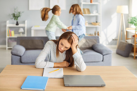 Young tired woman in noisy room, nanny, baby sitter single mother suffers from headache touching head, funny daughter and son jumping on sofa shouting, feels exhausted by noisy small kids at homeの写真素材