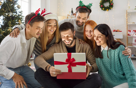 Group of happy friends gathering around their male mate who opening gift box. Joyful people sitting on floor, having party, celebrating Christmas together, exchanging presents. Merry Christmas conceptの写真素材