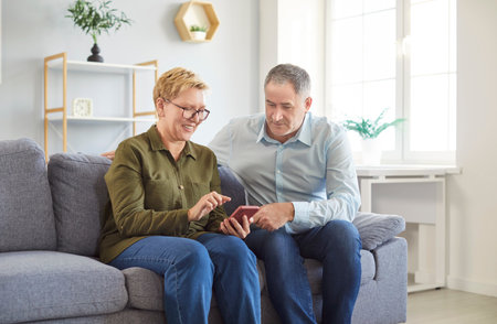 Happy elderly couple resting on sofa at home living room, enjoying leisure time together. They use a mobile phone for online entertainment, exploring the internet, and sharing joyful moments.の写真素材