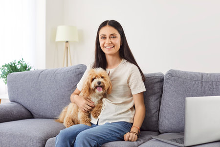 Cheerful dog owner young woman, cute girl stroking beloved pet on cozy home sofa, touching sweet doggy, looking at camera, smiling, enjoying weekend leisure with healthy adopted puppy friendの写真素材