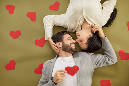 Top view portrait of a young man and woman looking at each other lying on green floor with red hearts and smiling close. Happy couple in love. Care, tenderness and Valentines day concept.の写真素材
