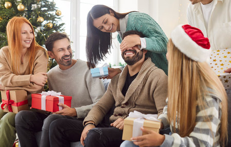 Portrait of a young woman congratulating man on Christmas holiday with present gift box sitting on sofa with friends at home. People celebrating New Year in decorated living room together.の写真素材