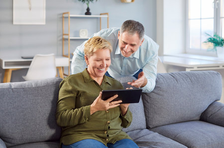 Senior happy couple at home, older man and woman having fun with computer tablet, elder holding mobile device, sitting on cozy couch at home, shopping online, reading good news in social networkの写真素材