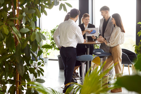 Business team gathered in office surrounded by greenery, discussing charts and financial results. Group of colleagues collaborating around table and brainstorming solutions in eco friendly workplace.の写真素材