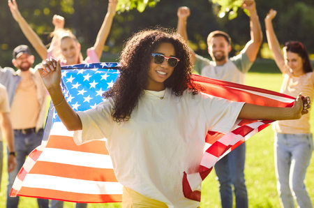 Portrait of happy african american woman holding usa flag in hands celebrating Independence Day standing in summer park outdoors with smiling people friends in background. Patriotism concept.の写真素材