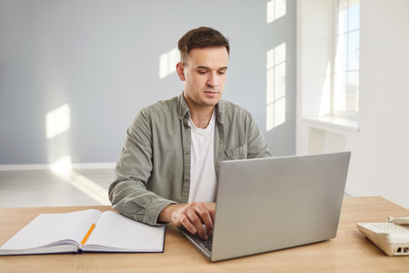 Confident young attractive business man looking seriously at computer monitor screen working at the desk on his workplace and typing in the office. Male person at work with laptop.の写真素材