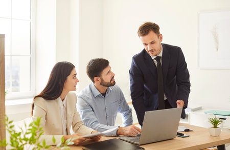 Group of three business colleagues working together in bright office. Young man in suit standing near desk, explaining something to seated man and woman who listening attentively. Teamwork concept.の写真素材