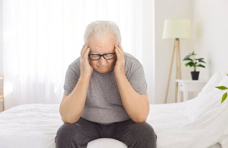 Sick elderly man is suffering from a headache or migraine while sitting on bed at home. Health problems, with the man feeling stressed, sad, and in pain, indicating his discomfort.の写真素材